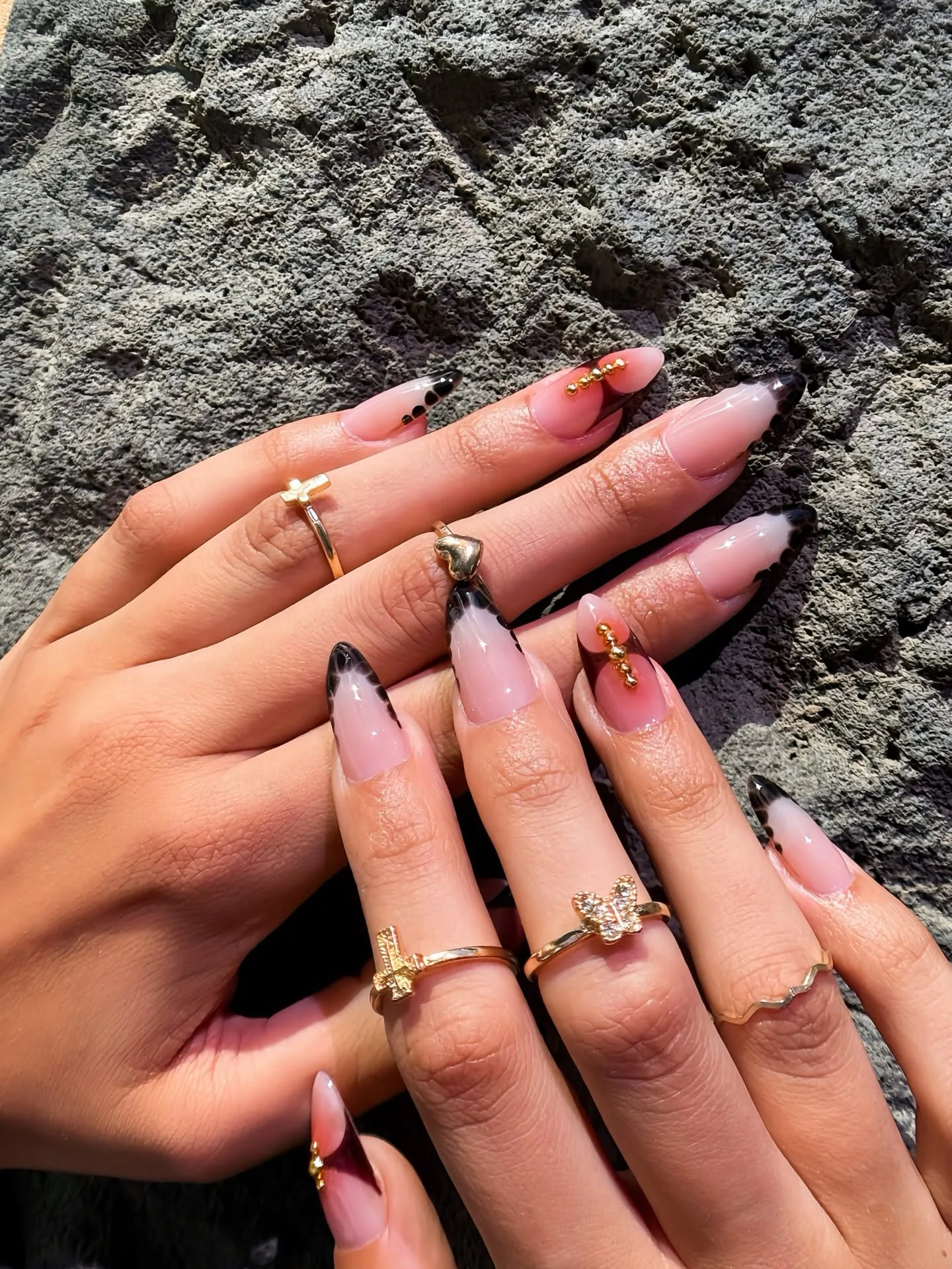 Black French-tip almond nails with gold micro-beads, hands resting on volcanic rock in sunlight