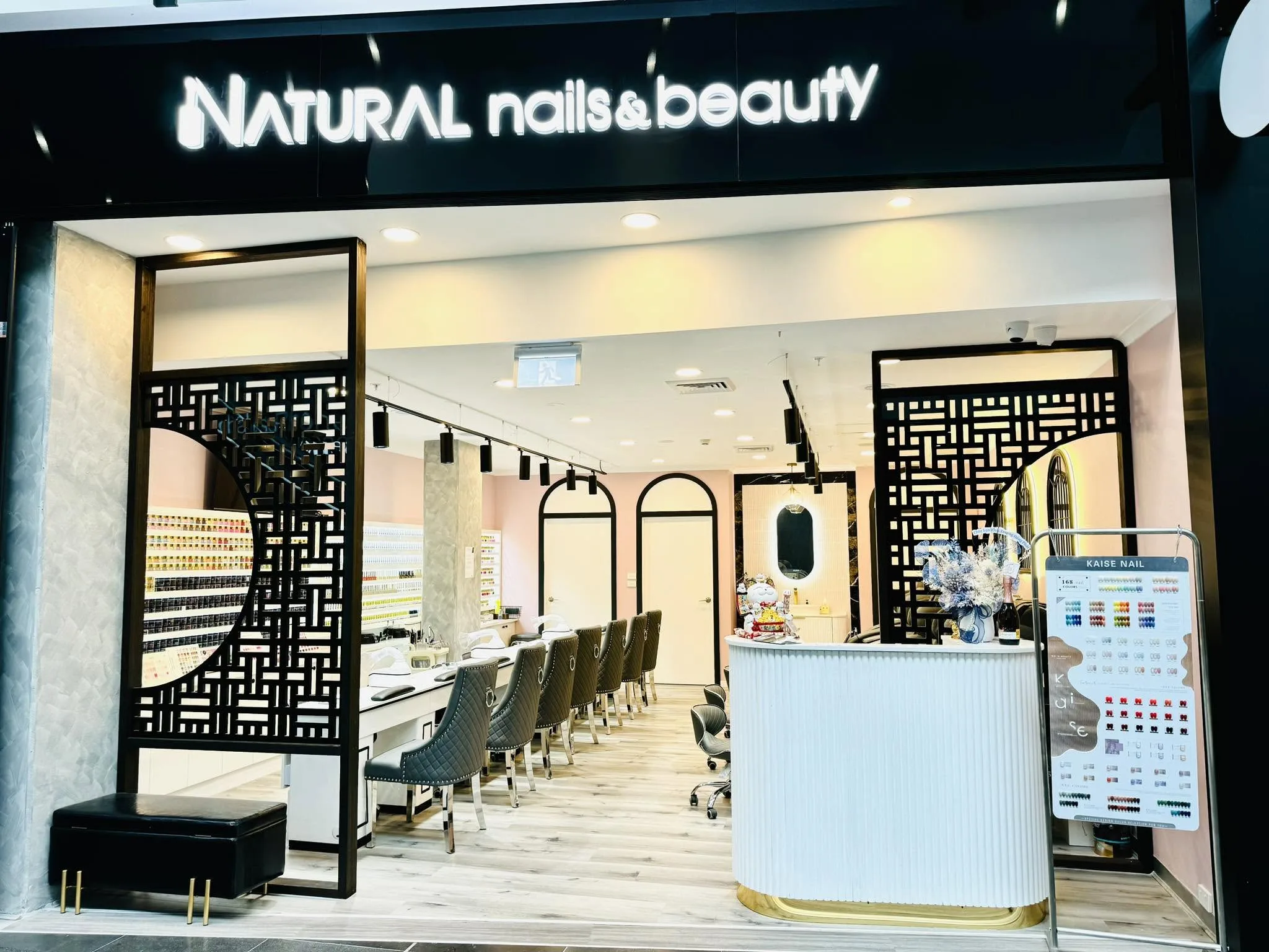 The Natural Nails & Beauty Coburg salon interior — lit shopfront sign, studded chairs, black carved-lattice screens, full Kaise Nail colour wall
